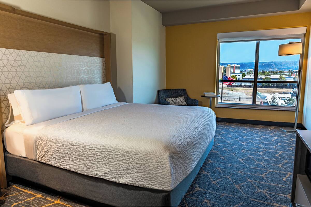 A hotel room with a large bed, a chair, a small table, and a window with a city view.