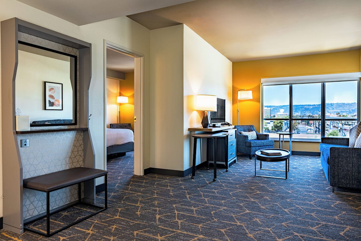 A hotel room with a bed, a desk, a chair, a sofa, and a large window offering a city view.