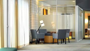 A modern hotel room with a glass partition, a round table with chairs, a floor lamp, and a green bench.