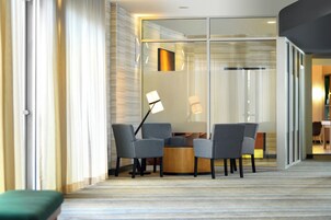 A modern hotel room with a glass partition, a round table with chairs, a floor lamp, and a green bench.