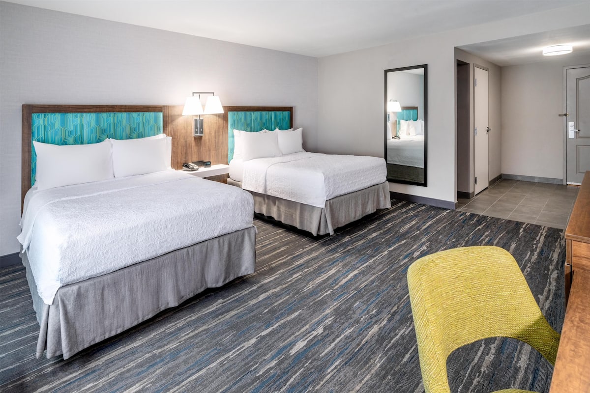 Quarto de hotel com duas camas, um espelho, uma cadeira e uma escrivaninha.