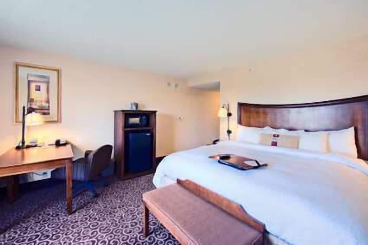 Quarto de hotel com uma cama, uma escrivaninha, uma cadeira, um pequeno armário e um quadro pendurado na parede.