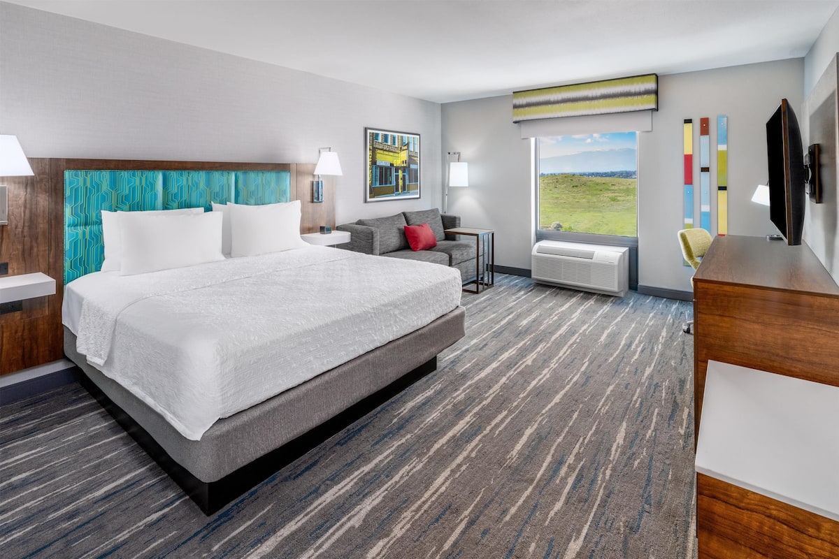 Quarto de hotel moderno com uma cama grande, um sofá, uma escrivaninha e uma janela com vista para o campo.