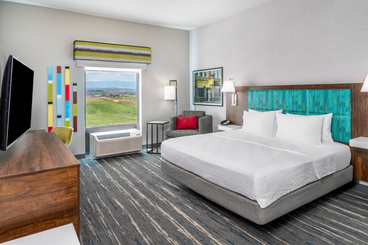 Quarto de hotel com uma cama grande, uma escrivaninha, uma cadeira, uma televisão, uma janela com vista para o campo e um quadro na parede.