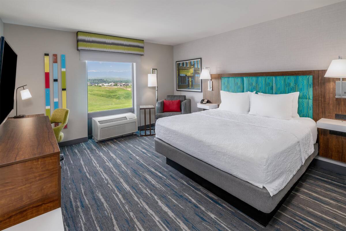 Quarto de hotel com uma cama grande, uma escrivaninha, uma cadeira, uma janela com vista e uma parede com um quadro abstrato colorido.