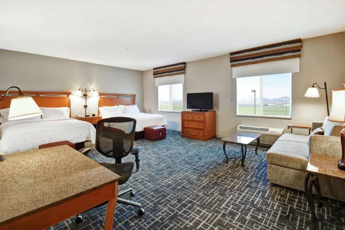 Habitación de hotel con dos camas, un escritorio, una silla, televisión, cómoda y grandes ventanales con vistas.