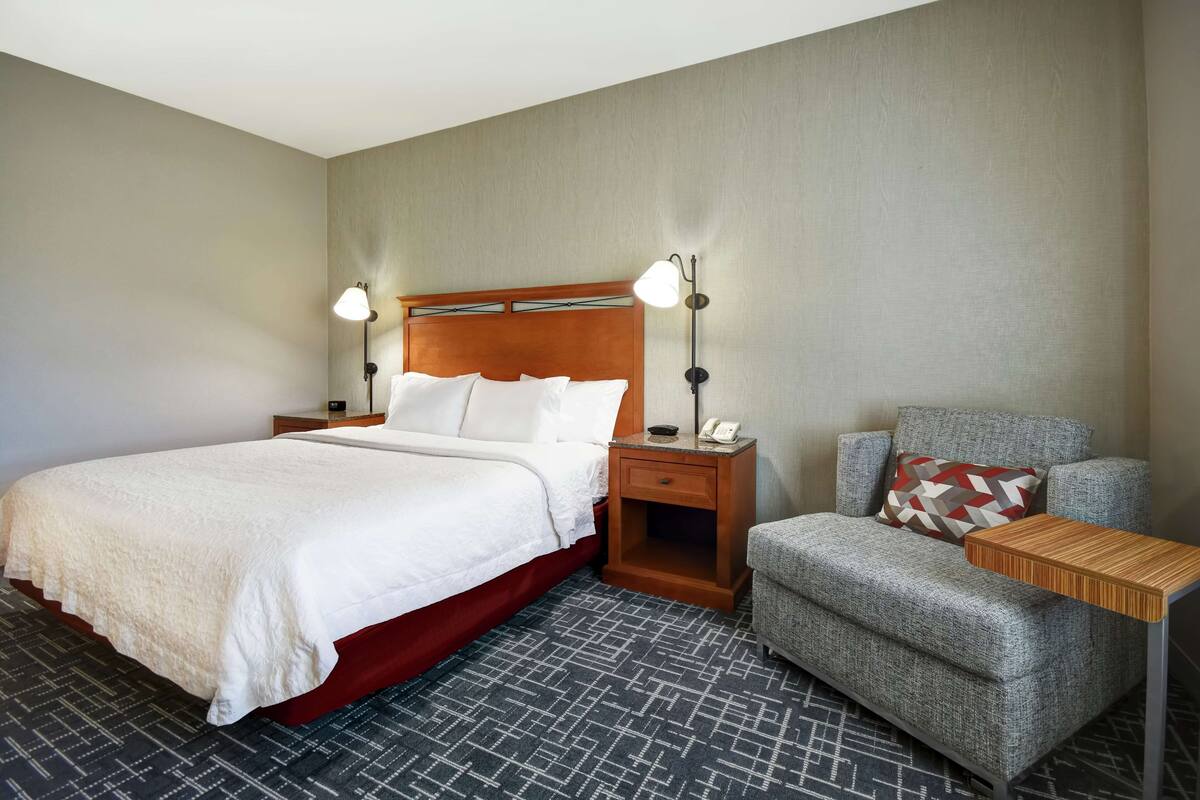 Una habitación de hotel con una cama, una mesita de noche con lámpara, una silla y una pequeña mesa.