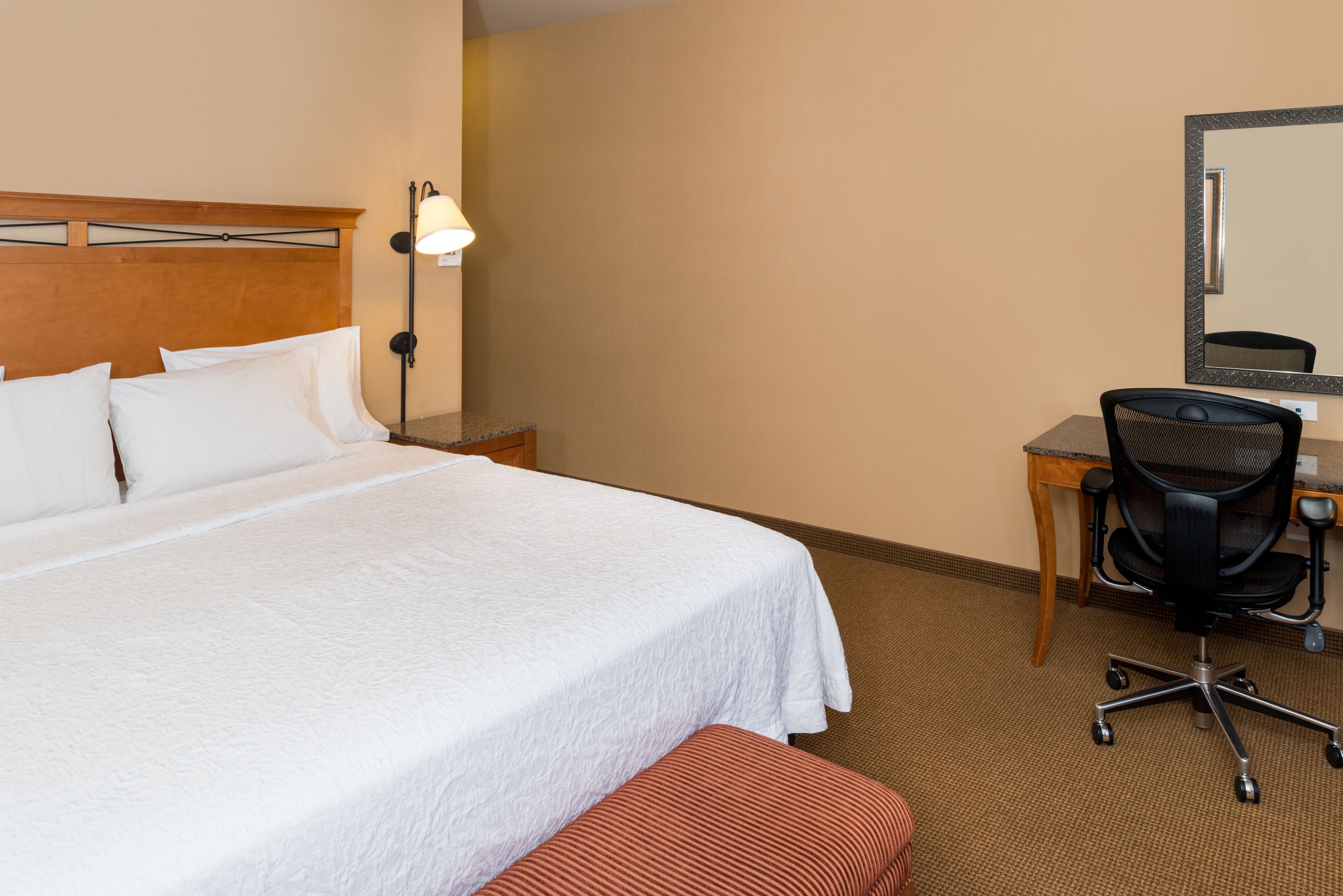A hotel room with a large bed, a desk with a chair, a lamp, and a mirror.