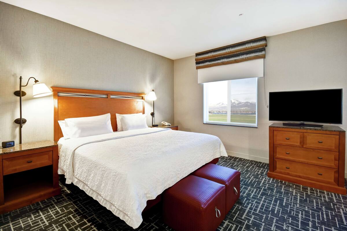 Habitación de hotel con una cama grande, televisión, mesita de noche de madera, lámpara, ventana con vistas y un banco rojo.