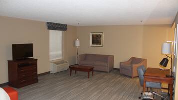 A hotel room with a bed, a wooden dresser, a television, a sofa, a desk with a chair, and a lamp.