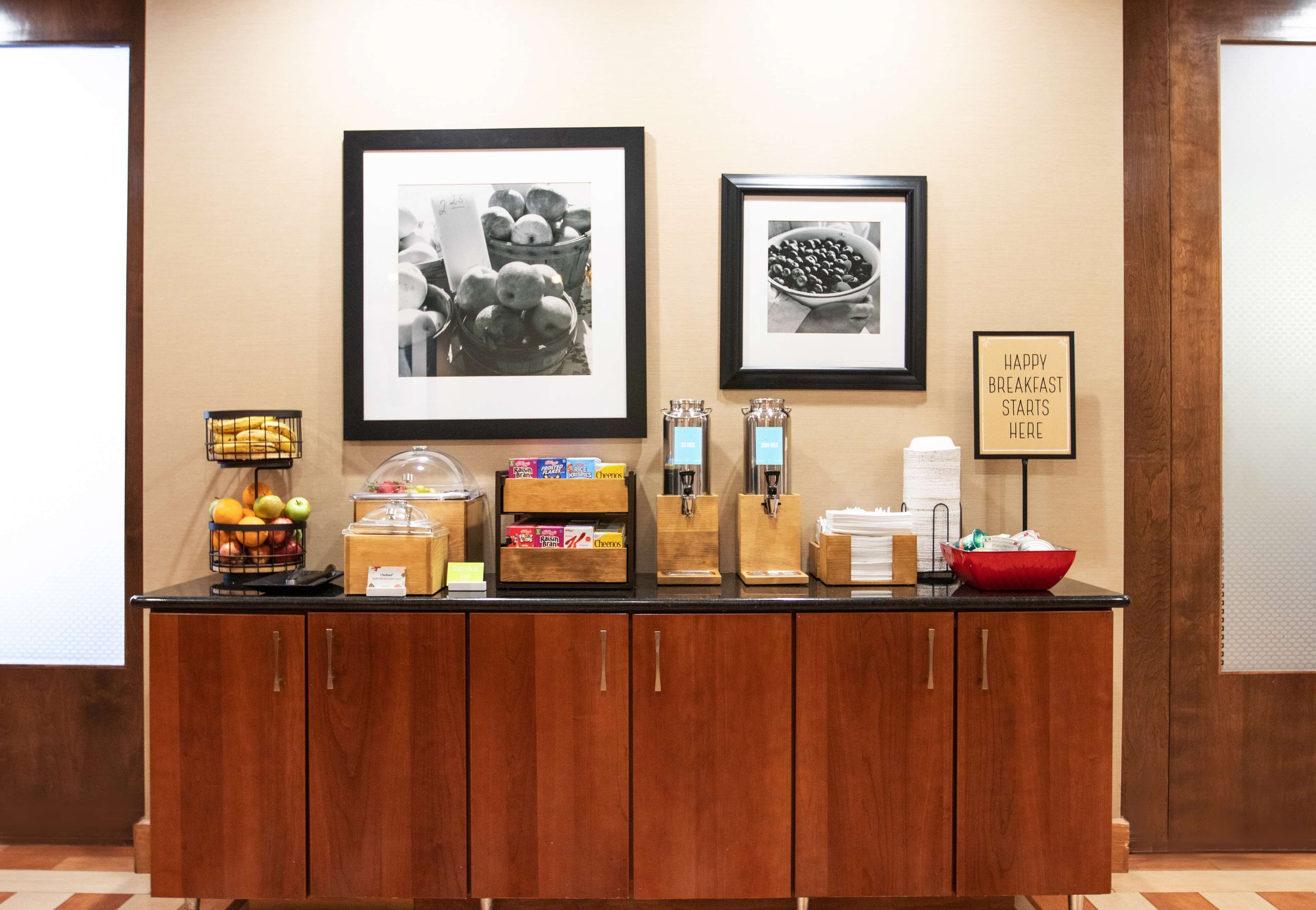 A breakfast bar with fruit, coffee, and tea dispensers, framed pictures, and a sign saying "Happy Breakfast Starts Here".