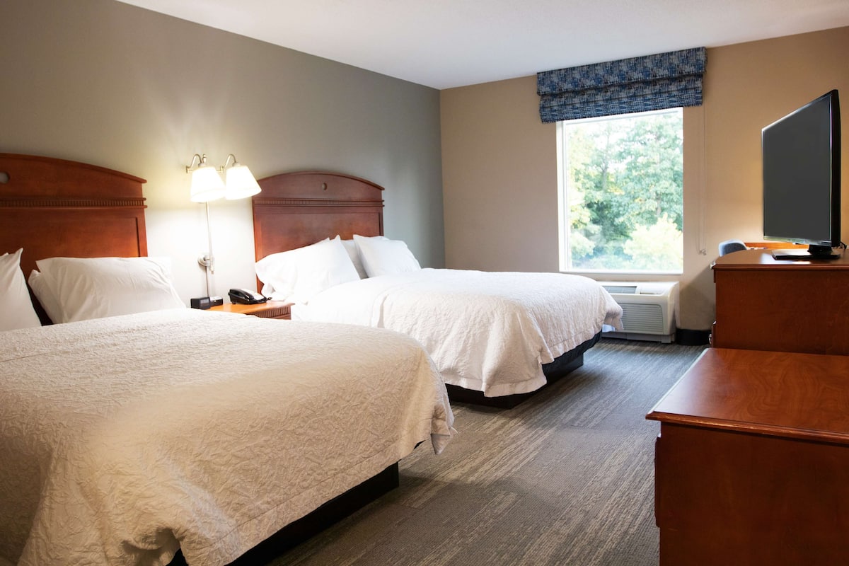 A hotel room with two beds, a desk, a television, and a window with a view of trees.