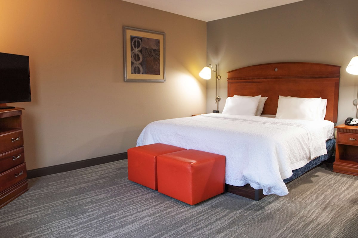 A hotel room with a large bed, two red ottomans, a wooden nightstand, a TV, and a framed picture on the wall.