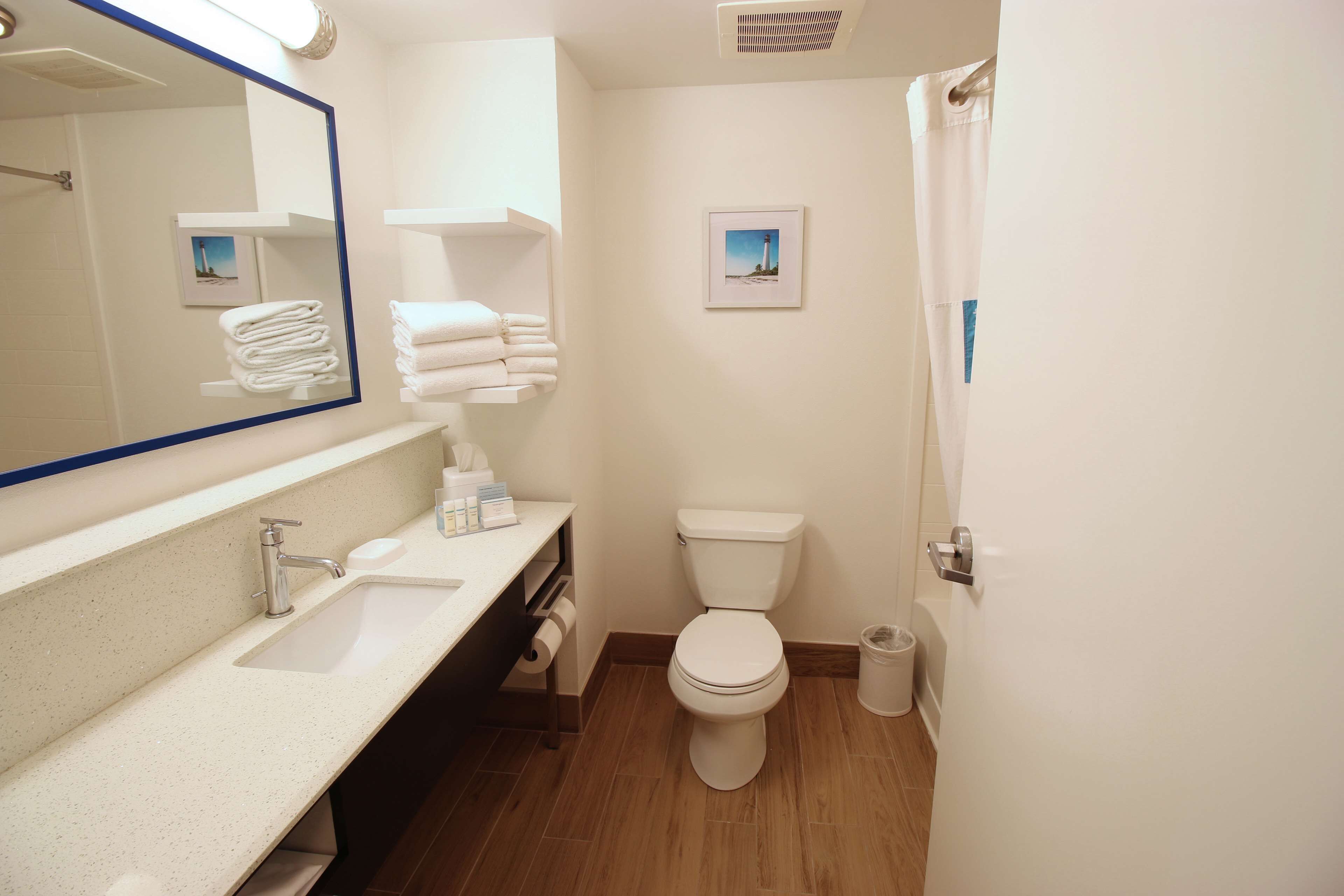 Un bagno con lavandino, servizi igienici e specchio. Asciugamani impilati su uno scaffale, e un quadro appeso alla parete.