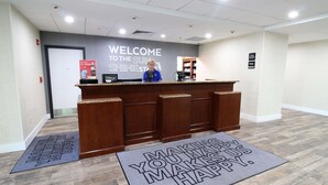 A reception desk with a 'Welcome to the Sun Shine State' sign, a 'Hilton' sign, and a 'Manager on Duty' sign. There are two doormats with motivational messages. - Hampton Inn & Suites Palm Coast (Palm Coast)