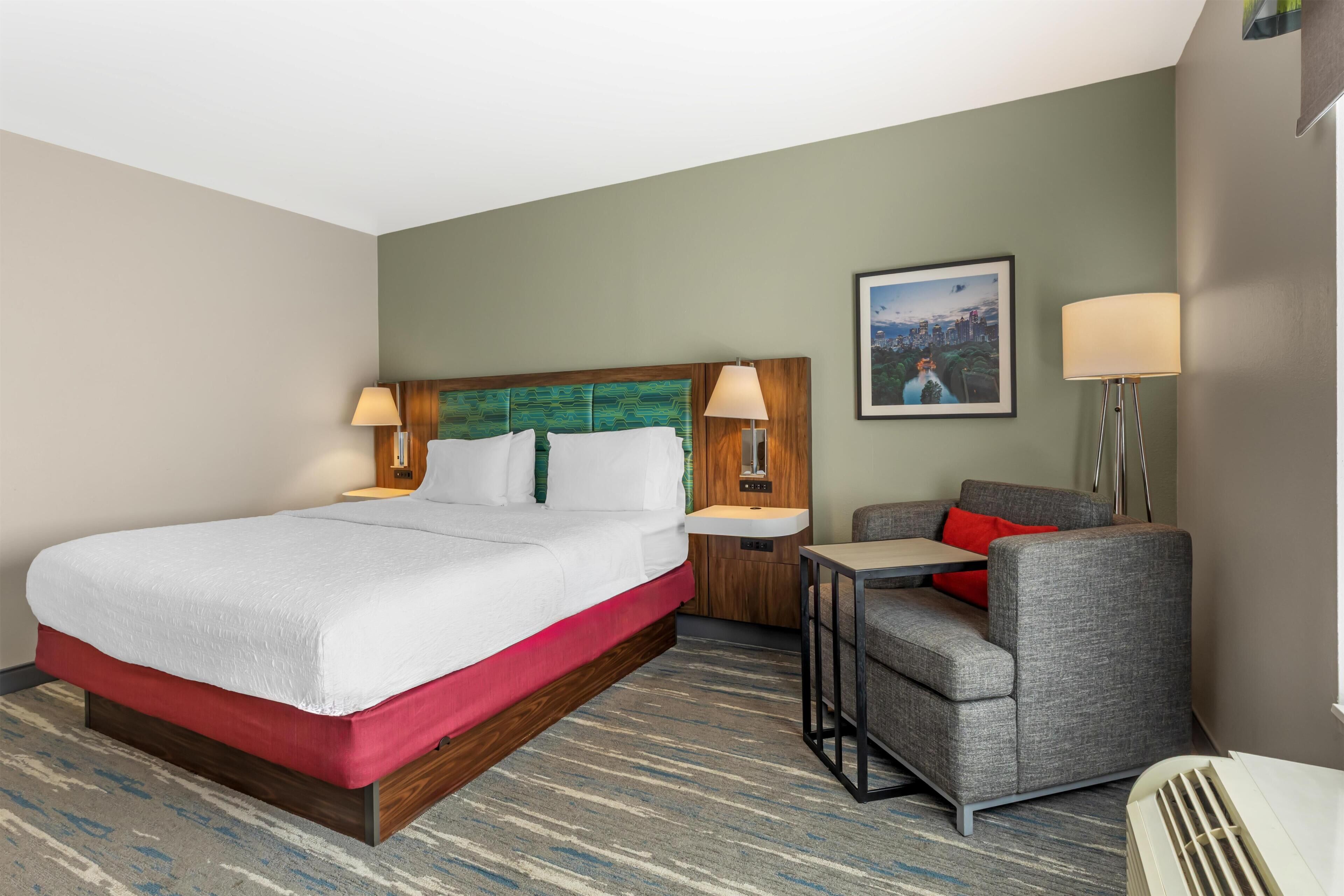 A hotel room with a bed, a desk, a chair, a nightstand, a lamp, and a painting on the wall.