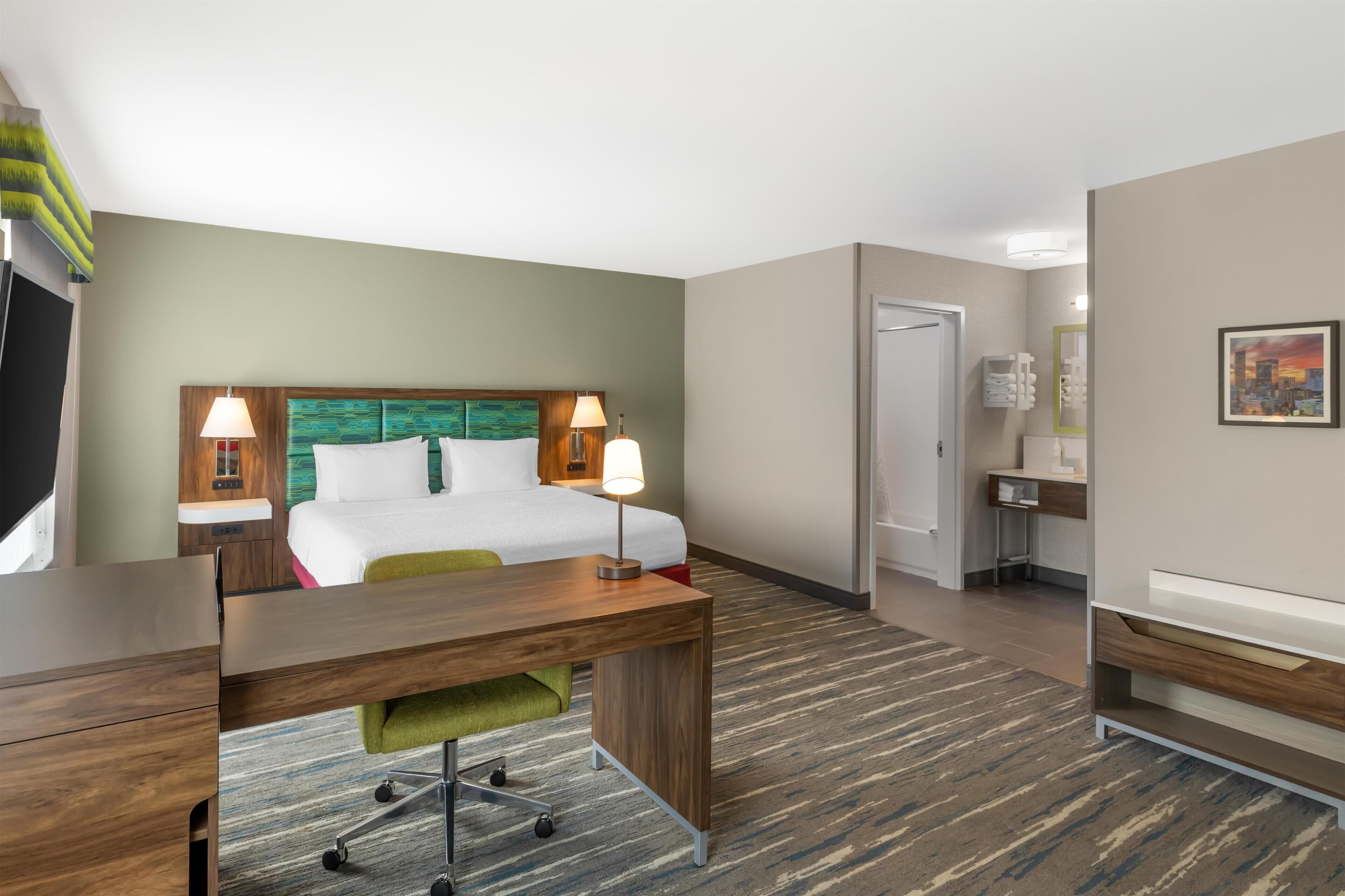 Quarto de hotel com cama, escrivaninha, cadeira e banheiro com pia e espelho.