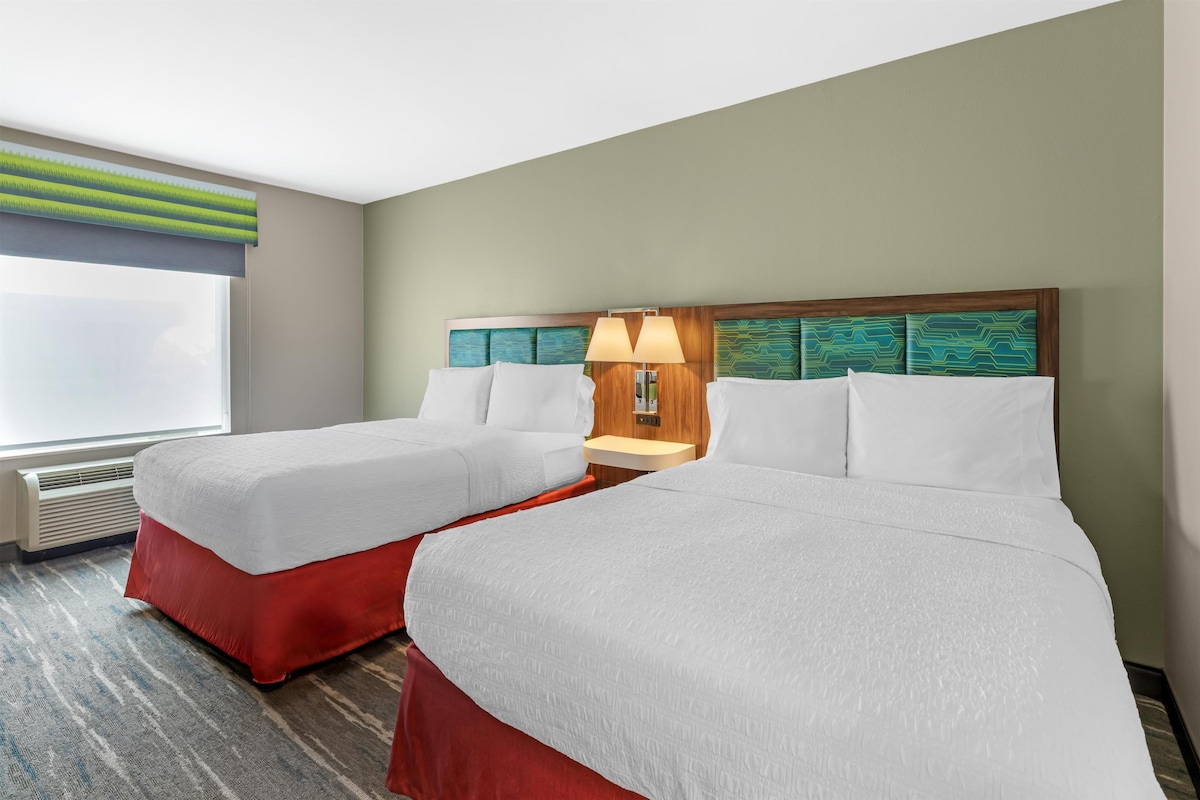 Una habitación de hotel con dos camas, una ventana con persianas, una mesita de noche con lámpara y un equipo de aire acondicionado de pared.