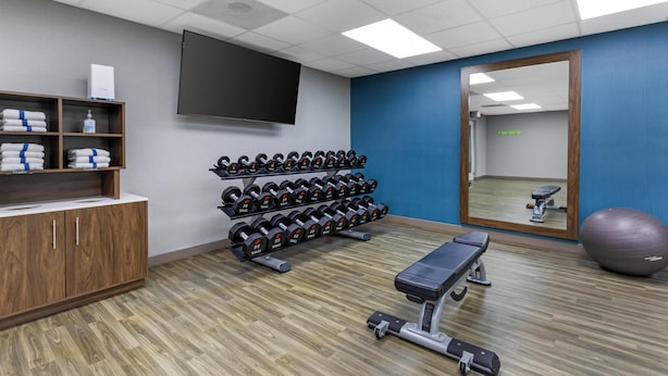 A gym with wooden flooring, a bench, a wall-mounted TV, and a rack of dumbbells.
