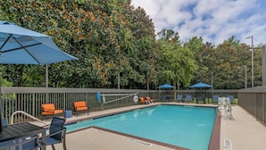 A swimming pool with lounge chairs and umbrellas, surrounded by a fence and trees.