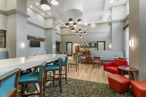 Un moderno vestíbulo de hotel con un bar central, zonas de estar y una cocina al fondo.