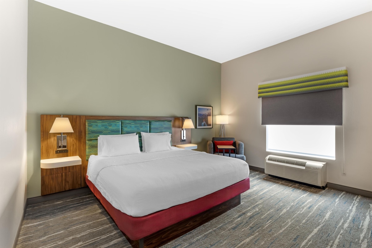 Habitación de hotel con cama, dos lámparas de noche, mesita auxiliar, ventana con persianas y aire acondicionado instalado en la pared.