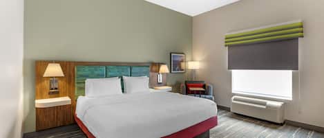 Quarto de hotel com cama, duas luminárias de cabeceira, mesa de apoio, janela com persianas e ar-condicionado fixado na parede.