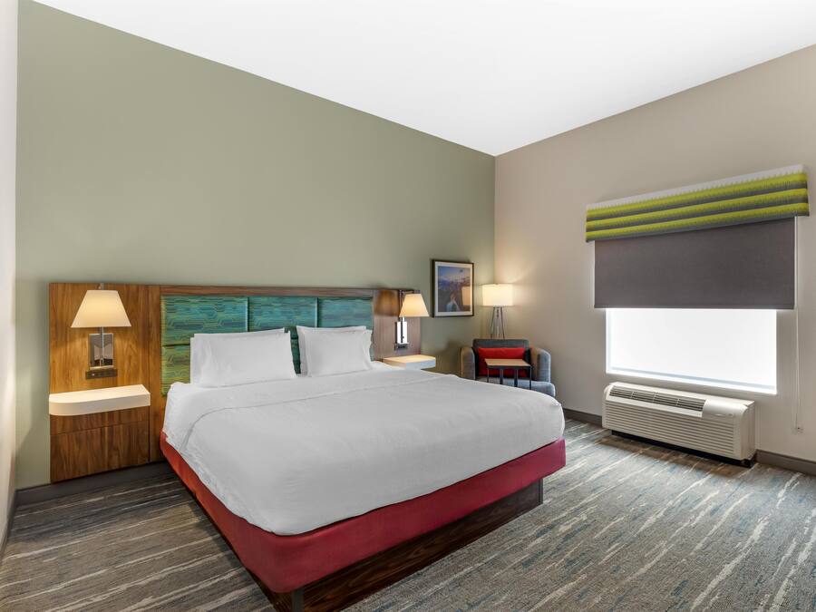 A hotel room with a bed, two bedside lamps, a nightstand, a window with blinds, and a wall-mounted air conditioner.