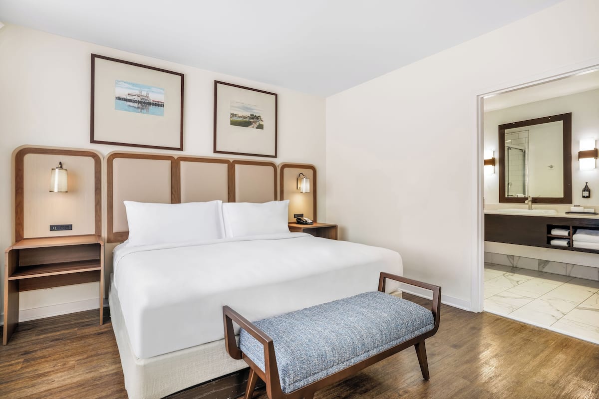 Ein Hotelzimmer mit einem großen Bett, Nachttischen mit Lampen, einer Bank, einem Badezimmer mit Spiegel und Waschbecken sowie zwei gerahmten Bildern an der Wand.