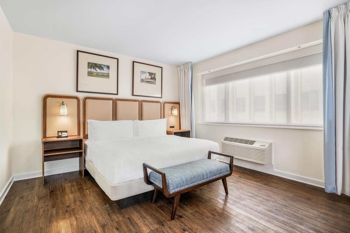 Ein Hotelzimmer mit einem großen Bett, Nachttischen mit Lampen, gerahmten Bildern an der Wand, einem Fenster mit Vorhängen und einer Bank am Bett.