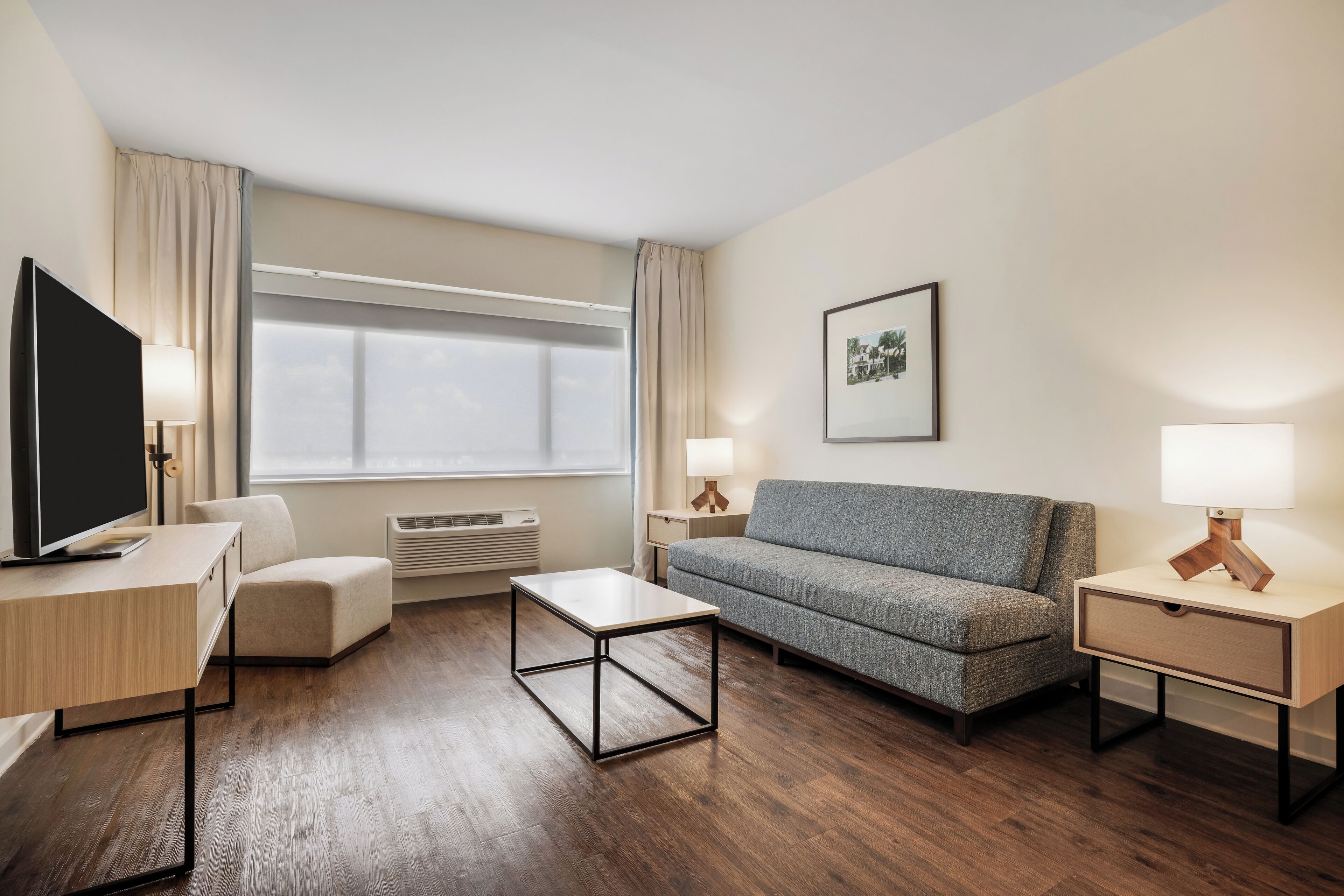 Una habitación de hotel moderna con suelo de madera, un sofá gris, una mesa de centro de madera, un televisor sobre un escritorio de madera y dos mesitas de noche con lámparas.