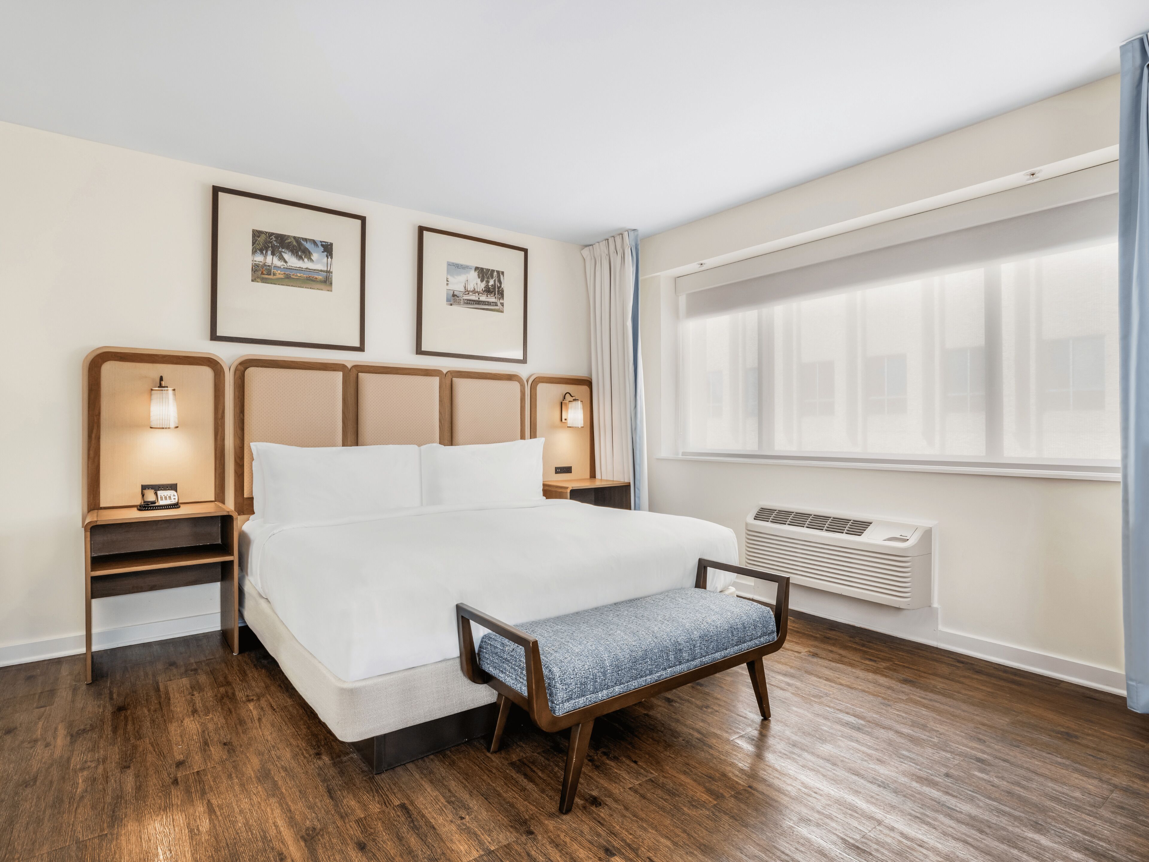 A hotel room with a large bed, wooden headboard, two bedside tables with lamps, a bench, a large window with curtains, and framed pictures on the wall.