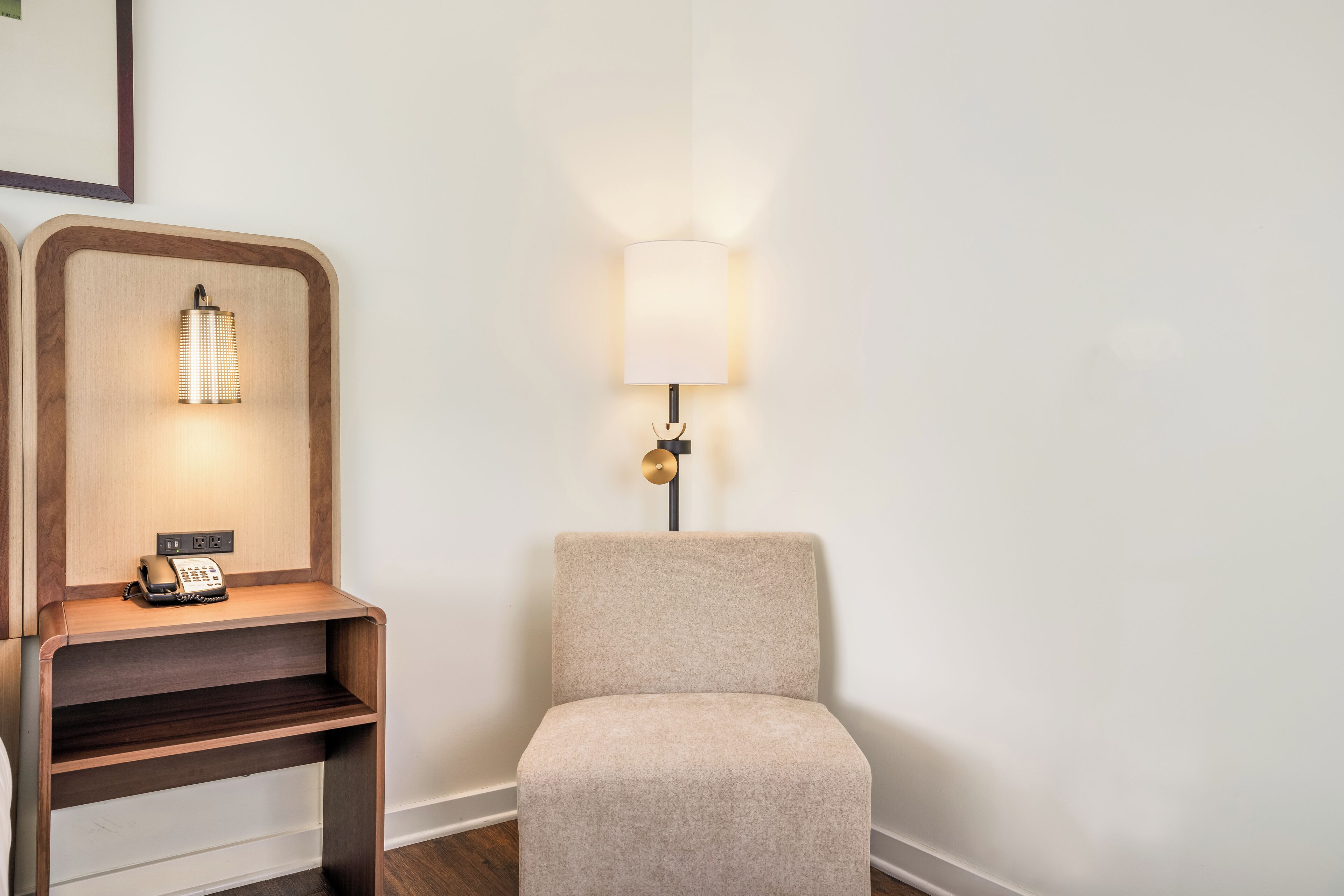 A hotel room with a beige armchair, a wooden nightstand with a telephone, and a wall-mounted lamp.