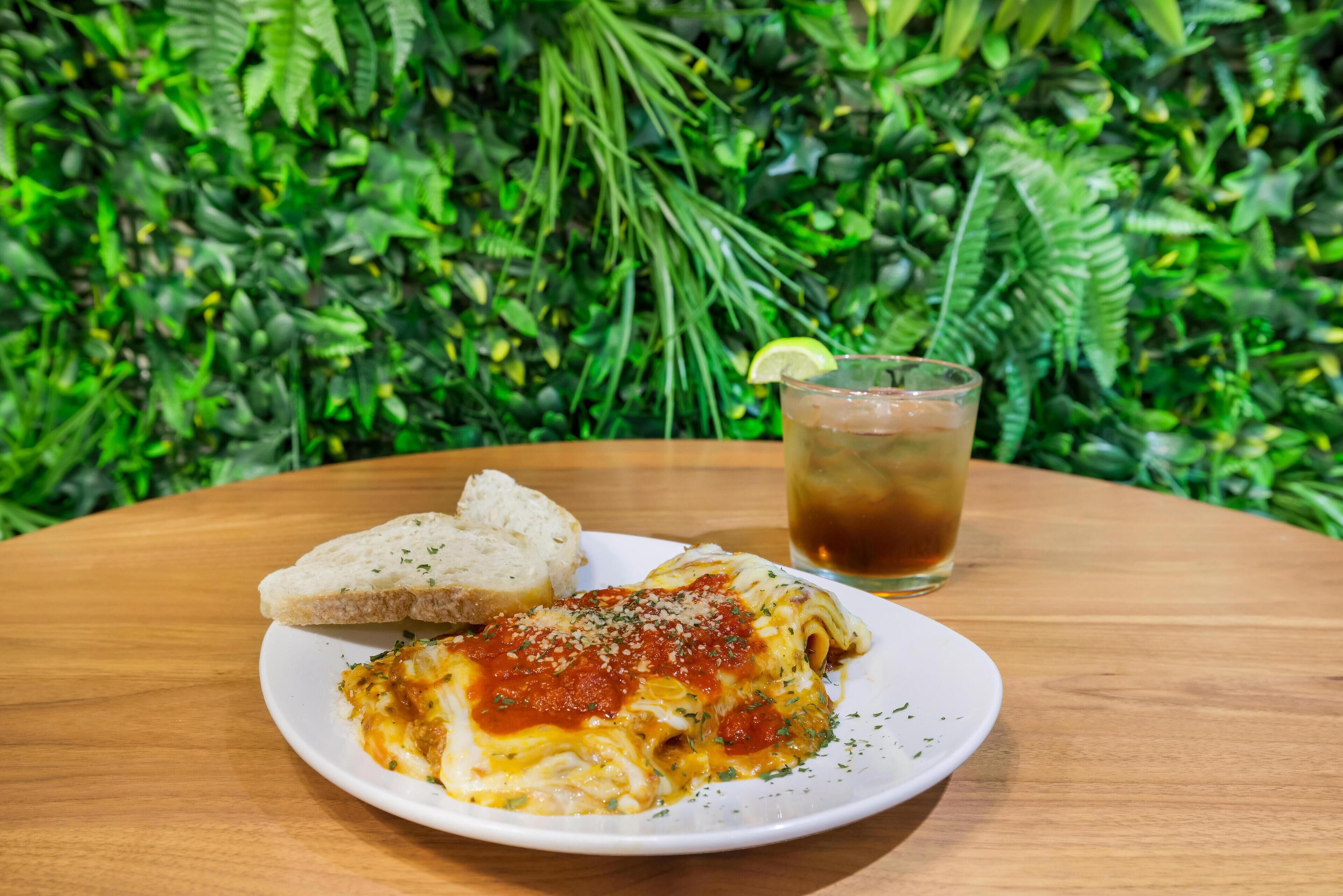 Lasagne med tilbehør af brød og et glas iste på et træbord med en grøn, løvfyldt baggrund.