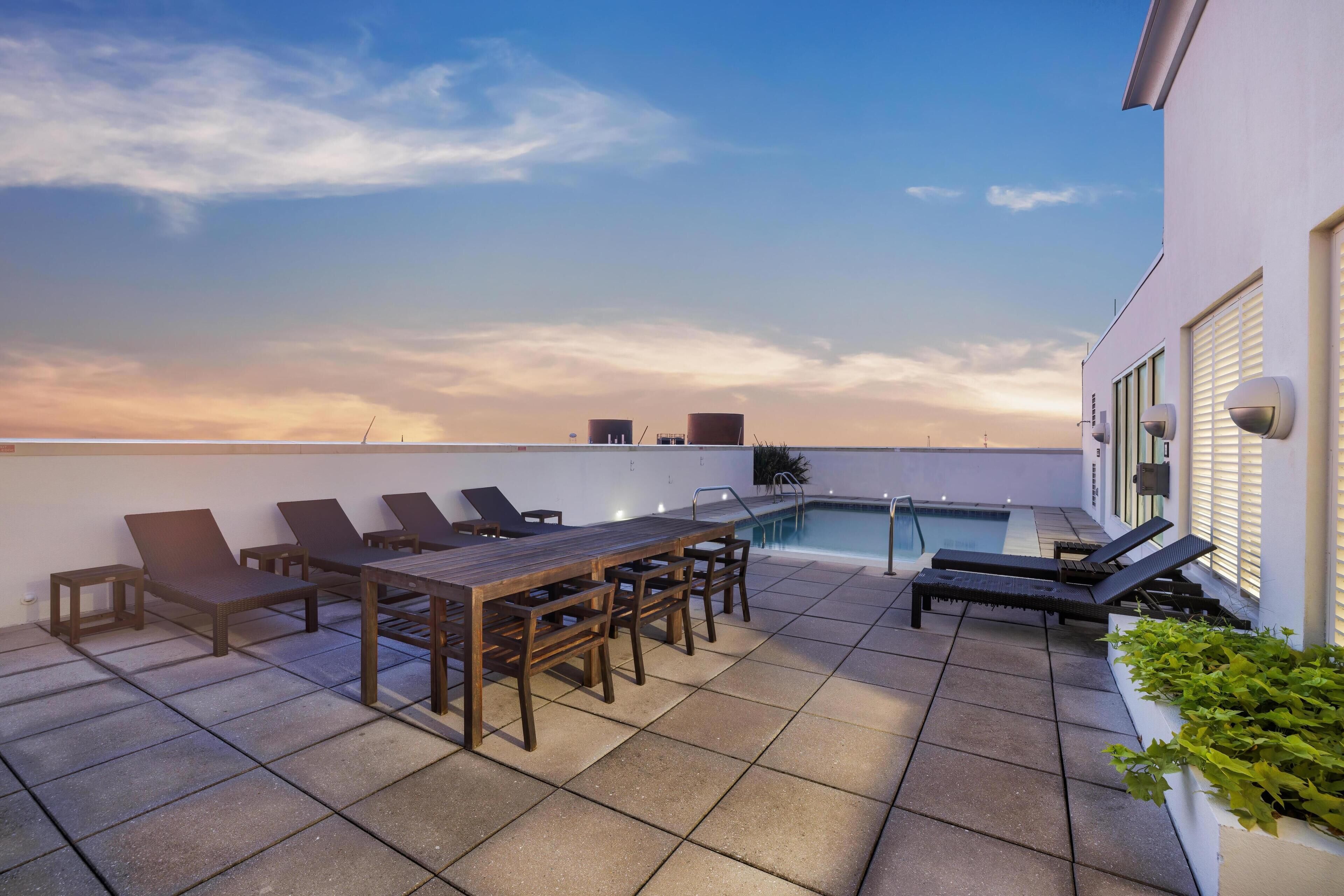 A rooftop terrace with a pool, lounge chairs, and a wooden table.