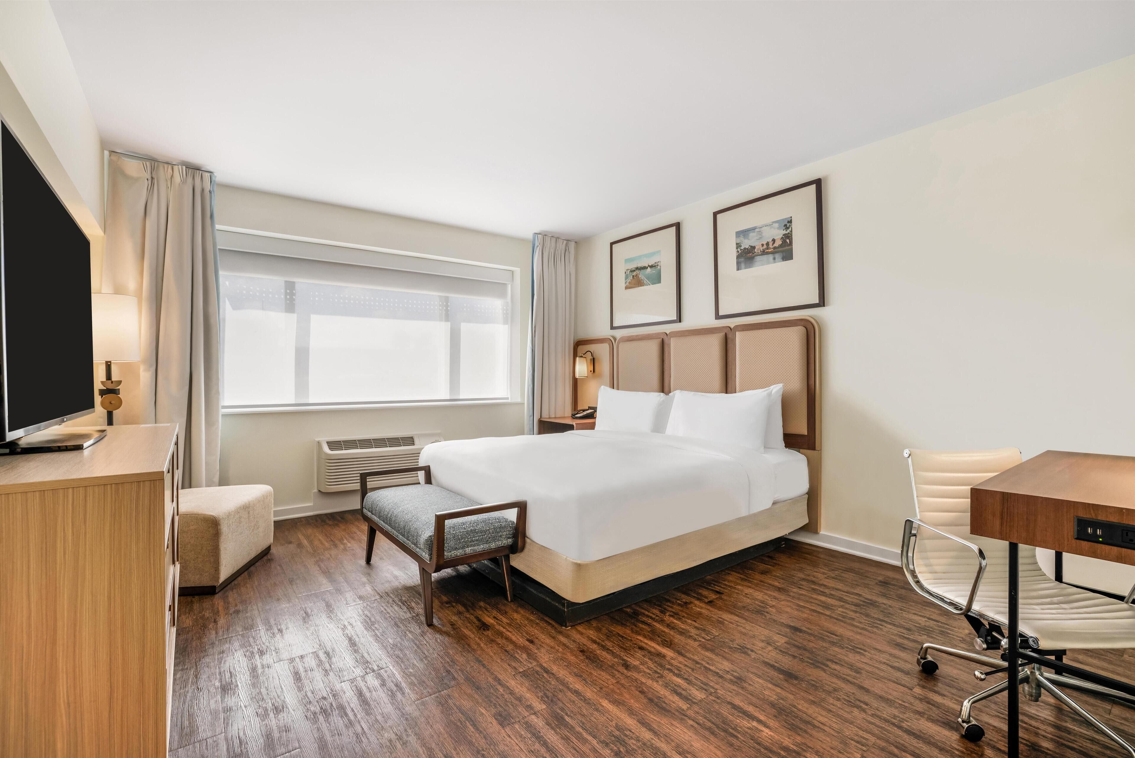 Ett hotellrum med en stor säng, ett skrivbord, en stol, en TV och ett fönster med gardiner.