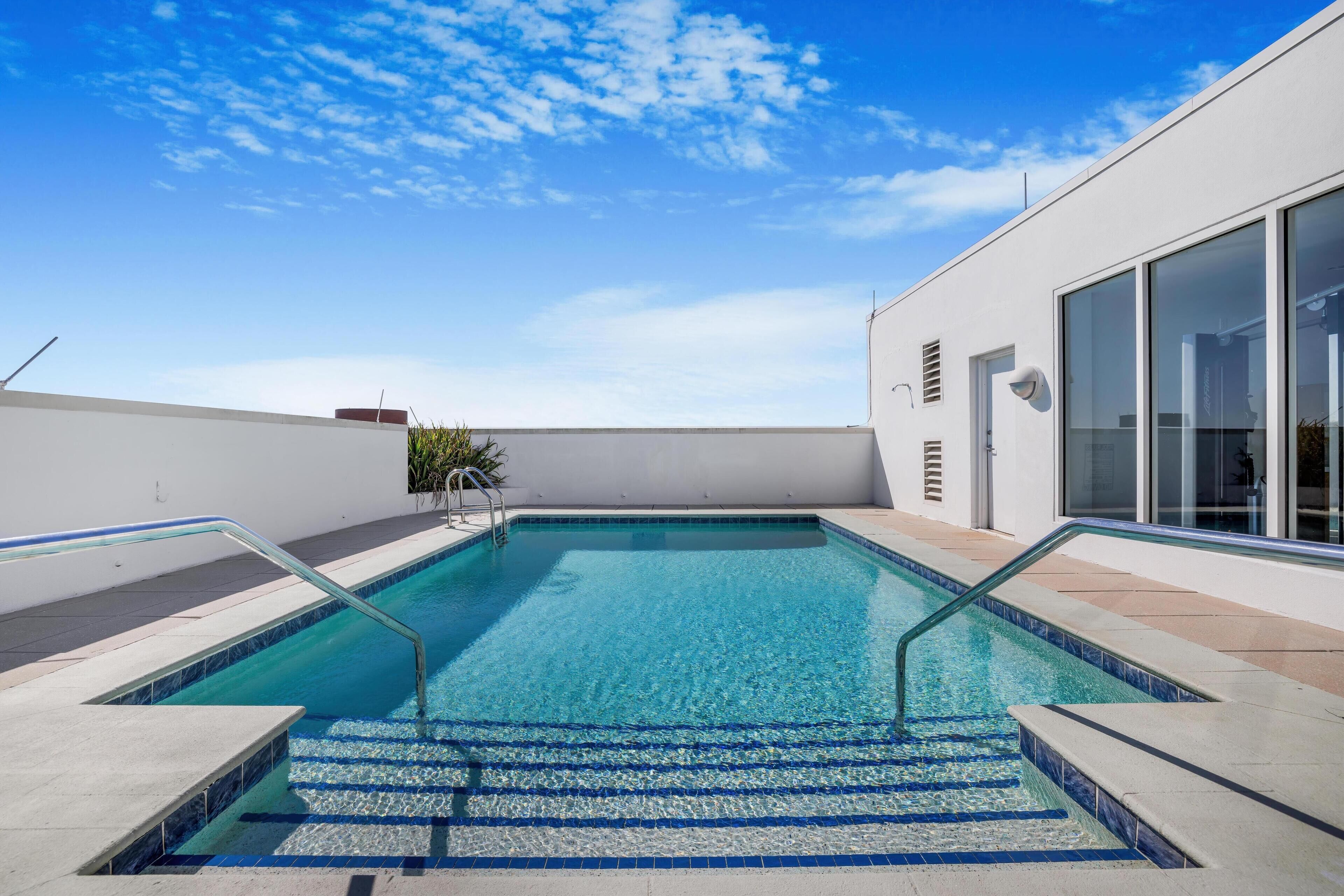 Una piscina en la azotea con agua azul cristalina, rodeada por un edificio blanco con grandes ventanales y un portal de vidrio.