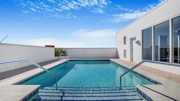 Una piscina en la azotea con agua azul cristalina, rodeada por un edificio blanco con grandes ventanales y un portal de vidrio.