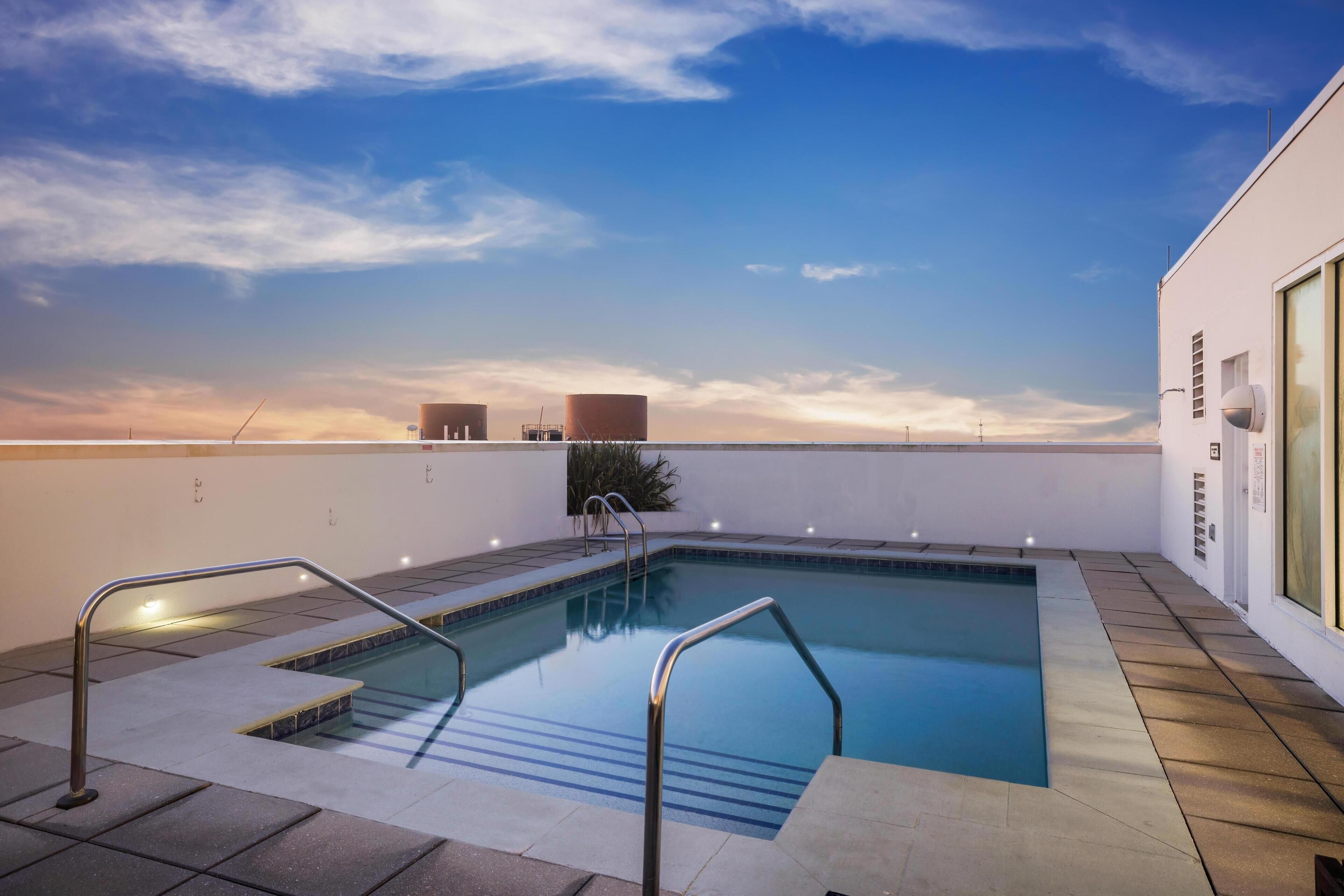 A rooftop swimming pool with clear blue water, metal railings, and a view of the sunset over the ocean.