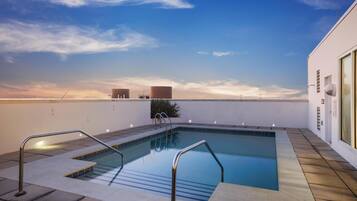 A rooftop swimming pool with clear blue water, metal railings, and a view of the sunset over the ocean.