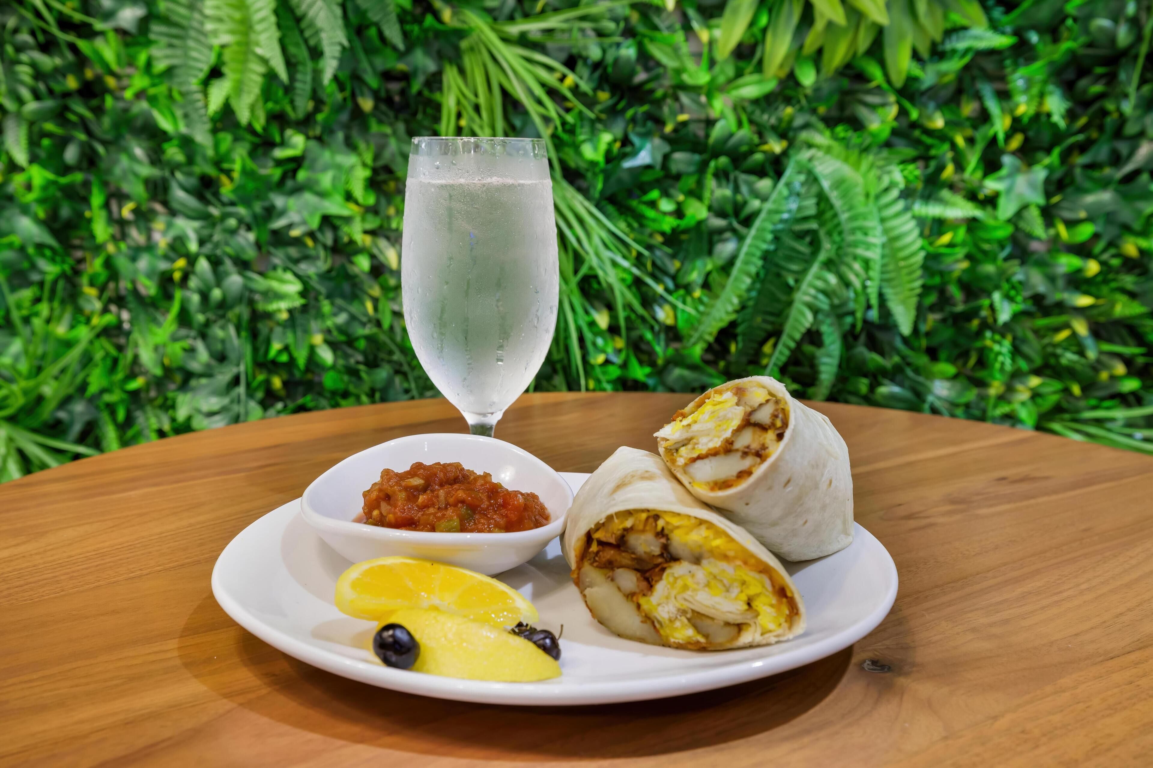 A plate with a wrap, a glass of water, a bowl of salsa, and lemon slices on a wooden table.