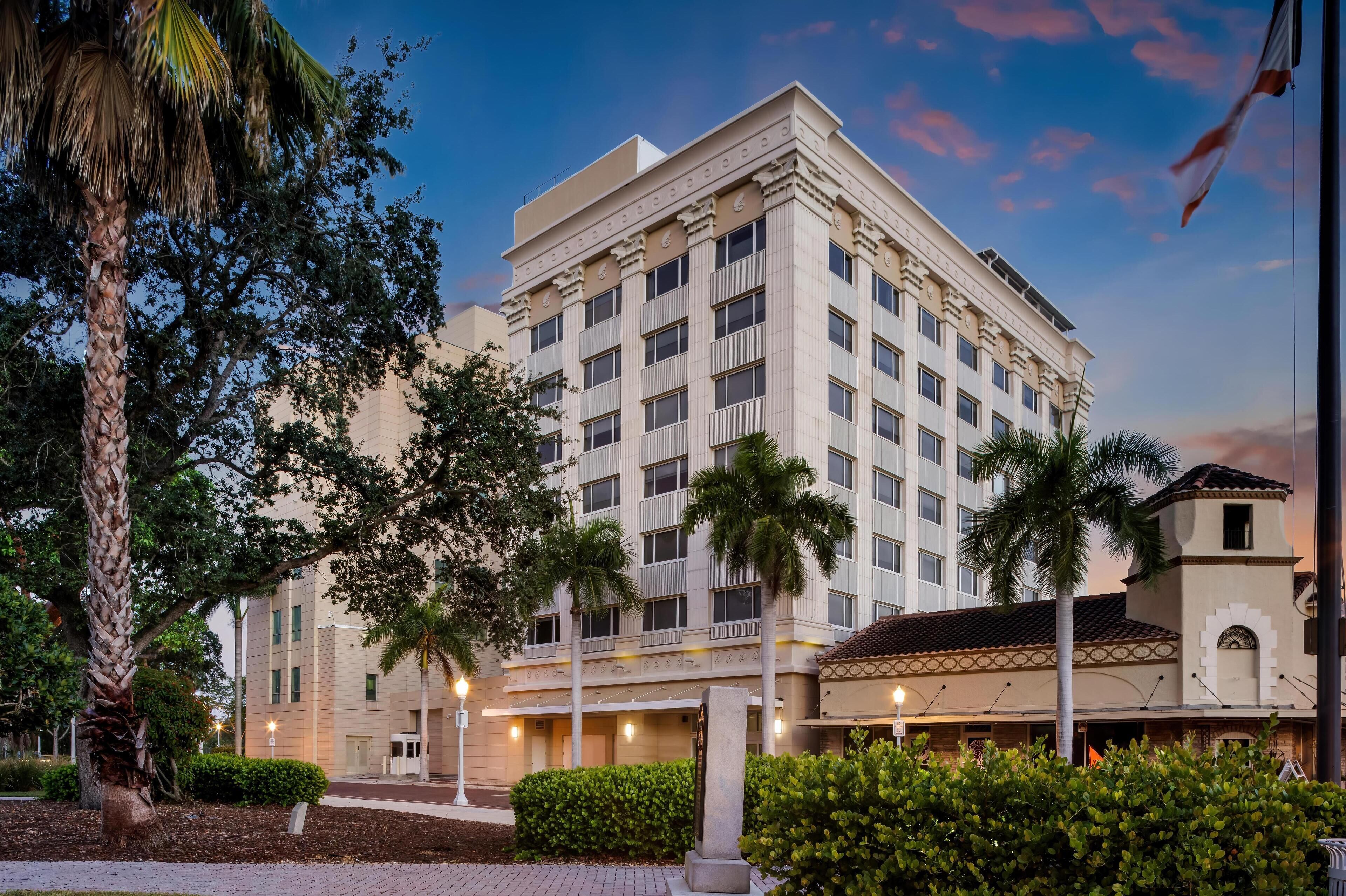 A multi-story hotel with a classic architectural design, featuring a central entrance, palm trees, and a well-maintained garden.