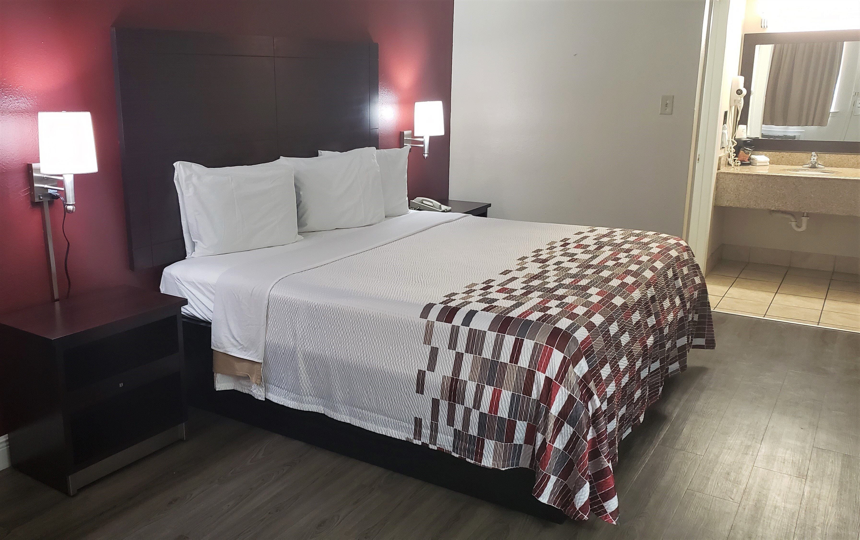 A hotel room with a bed, bedside tables, a nightstand, a wall-mounted lamp, a telephone, and a bathroom visible through an open door.