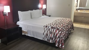 Hotellihuone, jossa on sänky, yöpöytä, sängyn vieressä oleva lamppu, puhelin ja kylpyhuone, joka näkyy avoimesta ovesta.