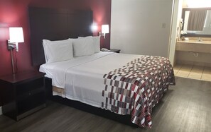 A hotel room with a bed, a nightstand, a bedside lamp, a telephone, and a bathroom visible through an open door. - Red Roof Inn Kenner – New Orleans Airport NE (Kenner)
