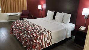 A hotel room with a large bed, a red chair, a desk with a phone, and a wall-mounted lamp. - Red Roof Inn Kenner – New Orleans Airport NE (Kenner)