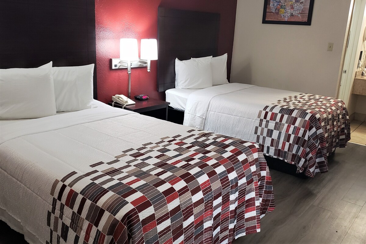 A hotel room with two beds, a nightstand with a phone and a clock, a red wall, and a bathroom with a mirror and towel rack.