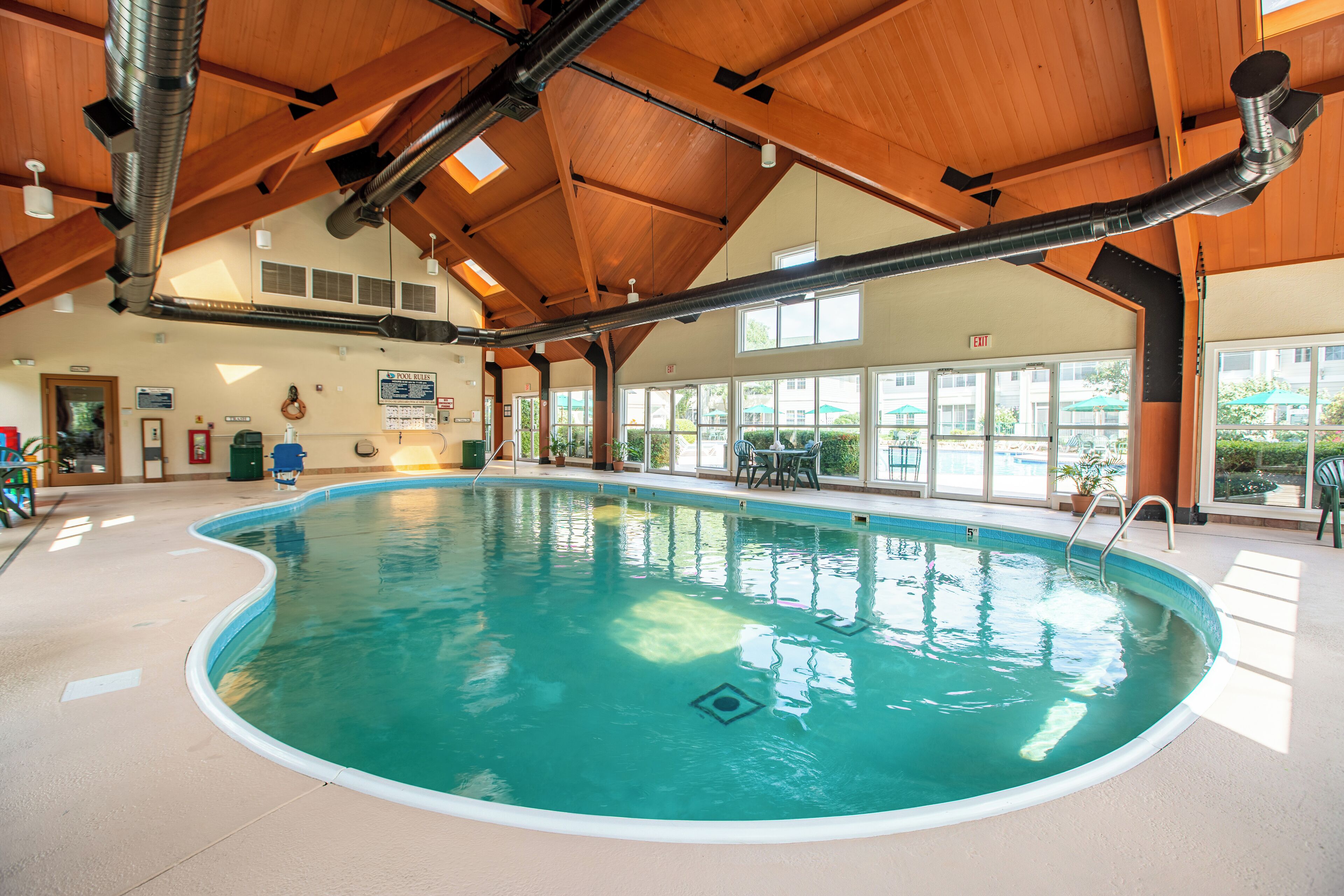 An indoor swimming pool with a wooden ceiling, clear windows, and a visible exit sign.