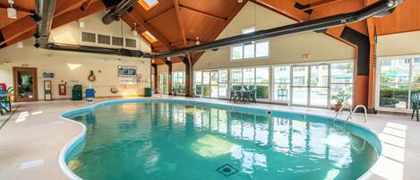 An indoor swimming pool with a wooden ceiling, clear windows, and a visible exit sign.