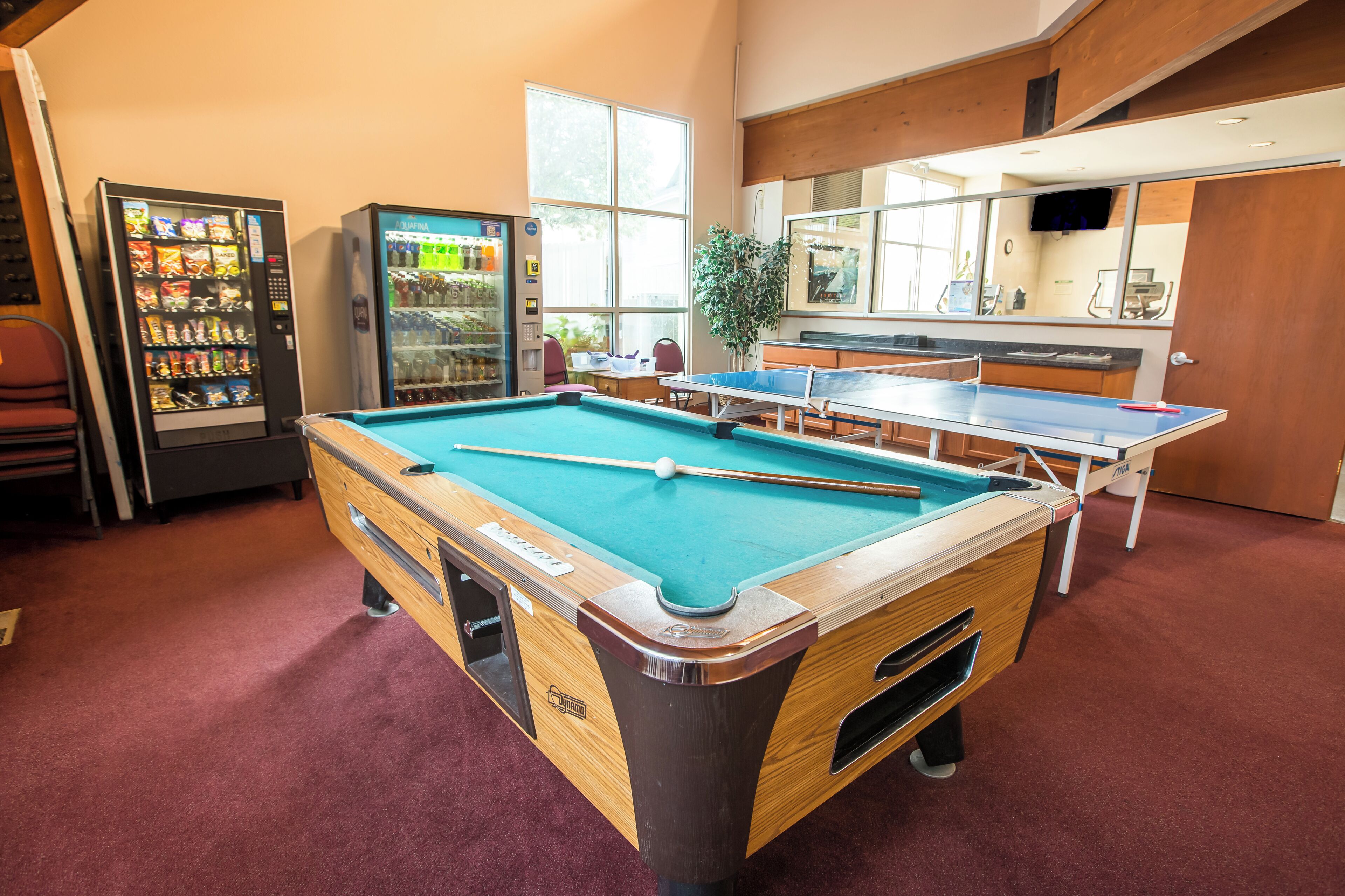 A pool table with a blue felt surface, a ping pong table, and vending machines in a room with red carpet flooring.